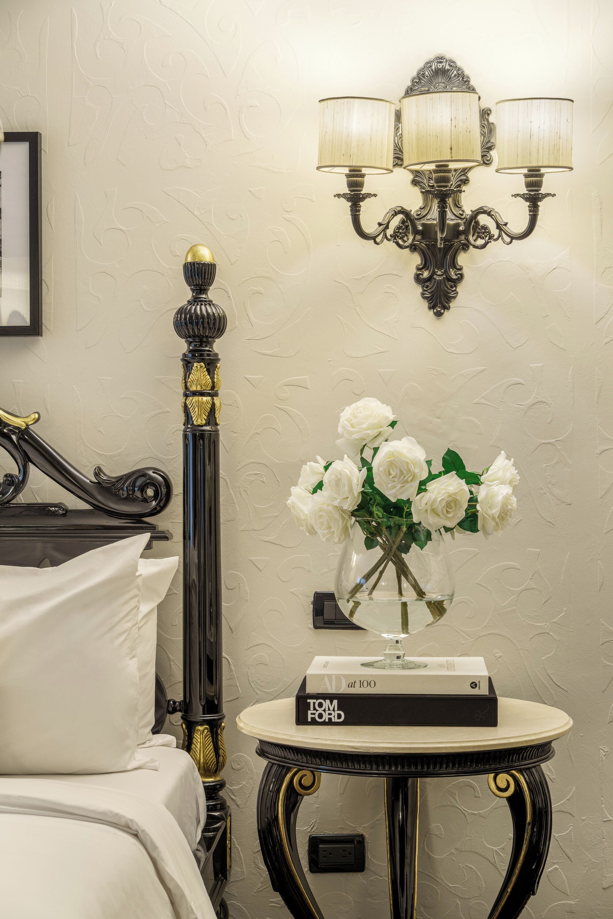 A hotel room with a bed, a nightstand with a book, a vase with flowers, and a wall-mounted lamp.