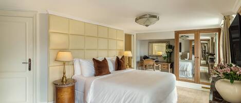 A spacious bedroom with a large bed, a bedside table with lamps, a mirror, and a view into the adjacent room.
