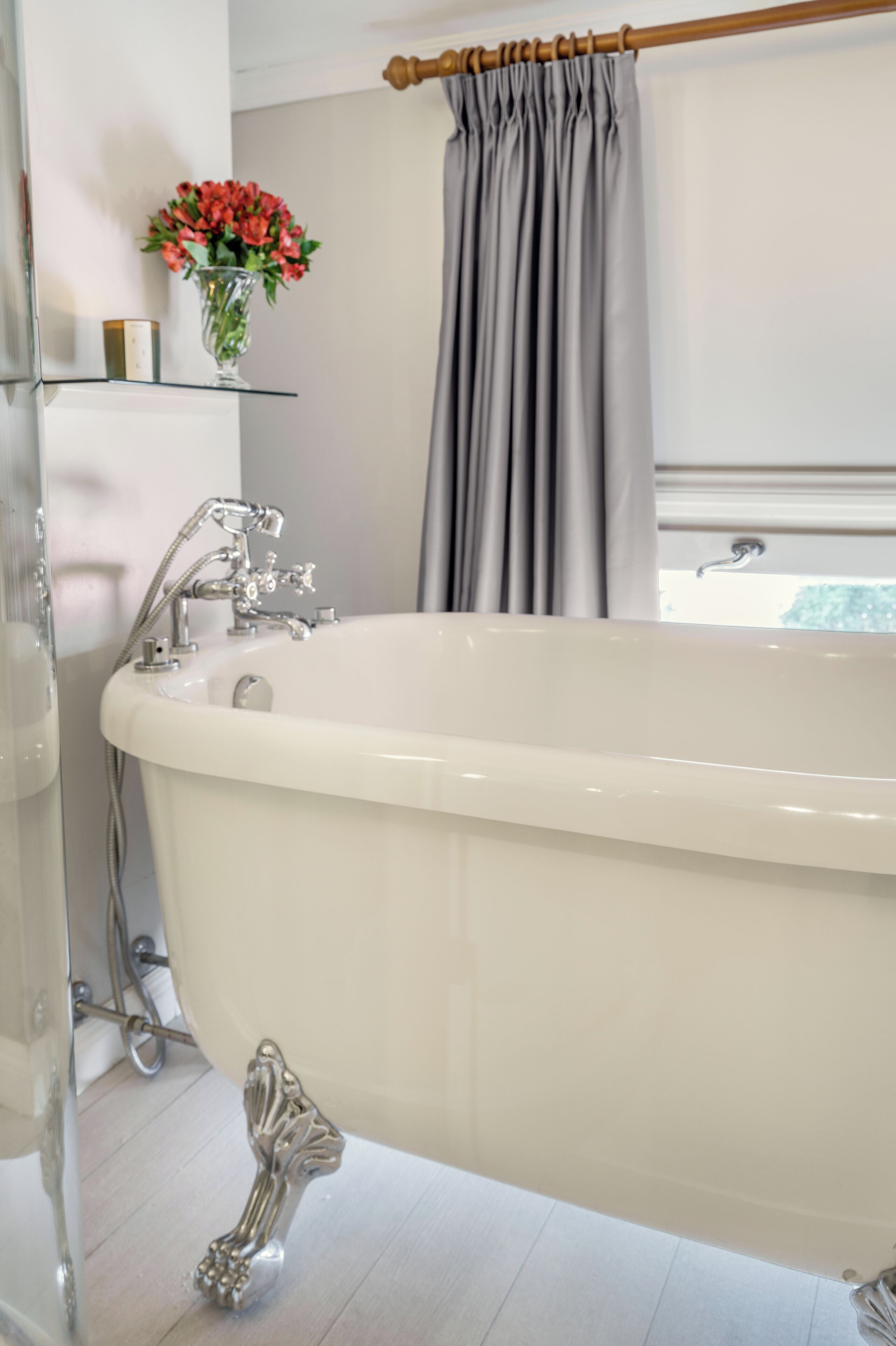 A bathroom with a freestanding bathtub, a glass shelf with a vase of flowers, and a window with curtains.