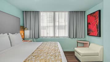 A hotel room with a large bed, a bedside table with a lamp, a small sofa, a window with curtains, and a wall-mounted artwork.