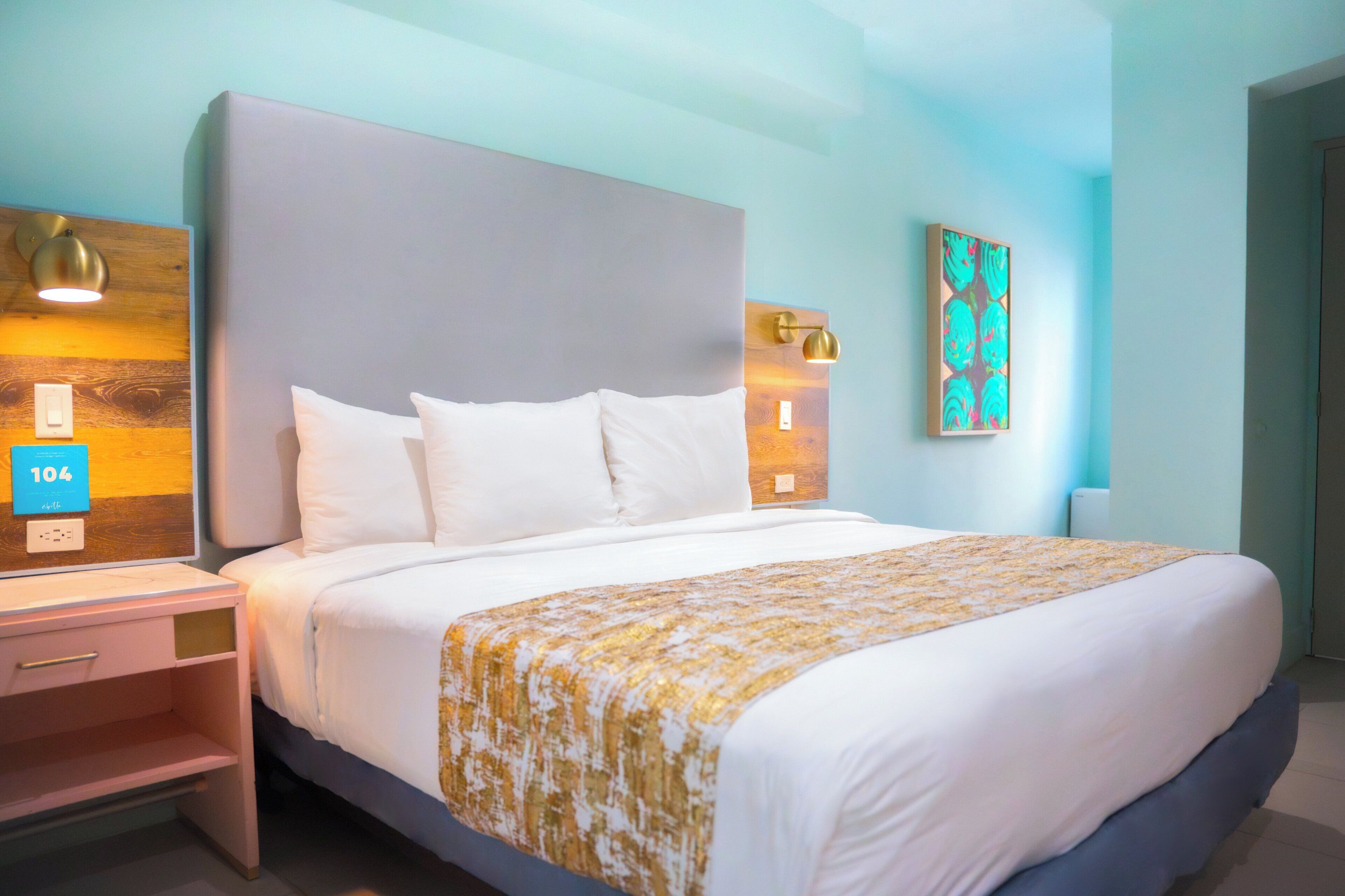 Una cama bien hecha con ropa blanca y un plaid estampado en dorado, una mesita de noche con una lámpara, y una lámpara fijada a la pared.