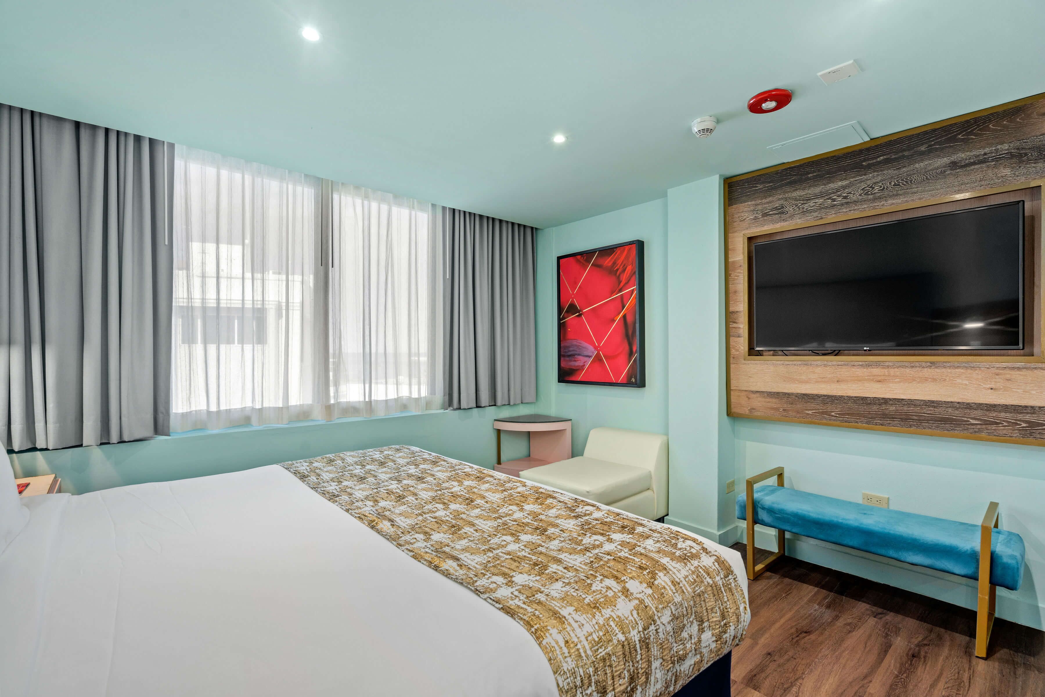 Une chambre d’hôtel avec un grand lit, une petite table de chevet, une chaise, un banc, un téléviseur fixé au mur et une fenêtre avec des rideaux.