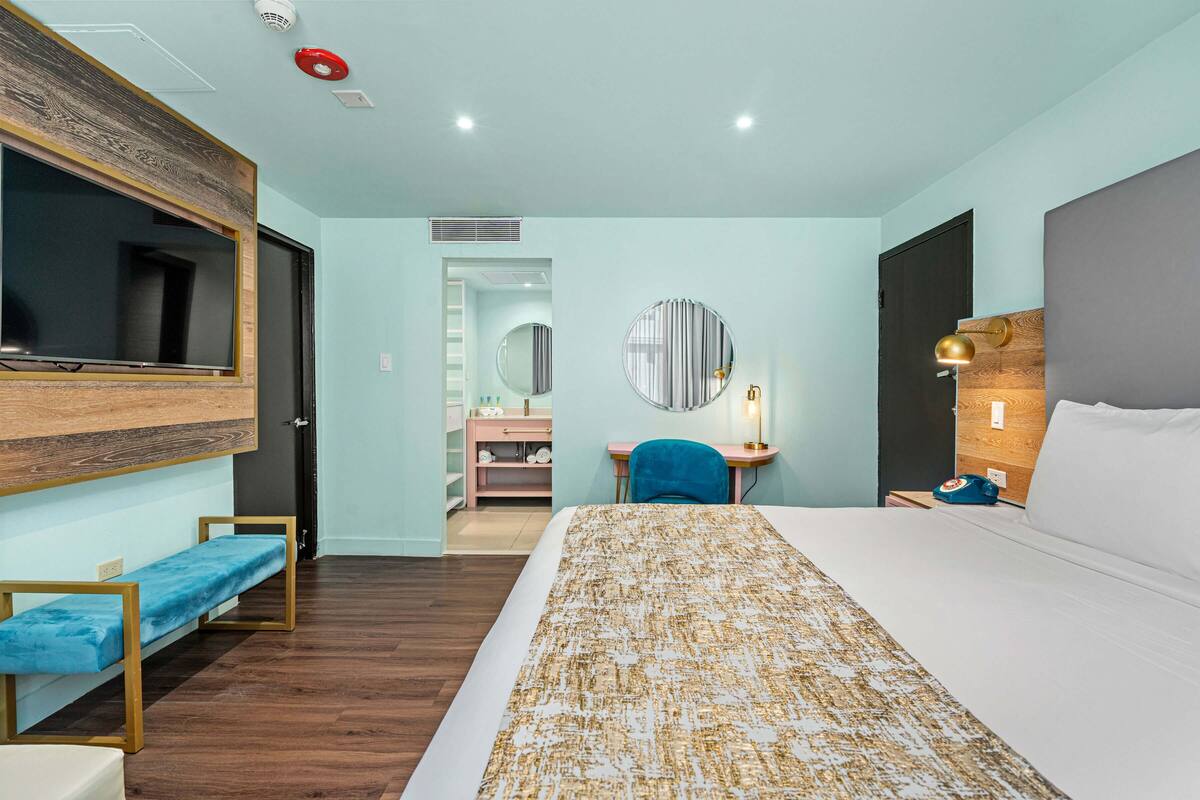 A hotel room with a bed, a desk, a chair, a TV, a mirror, and a bedside table with a lamp.