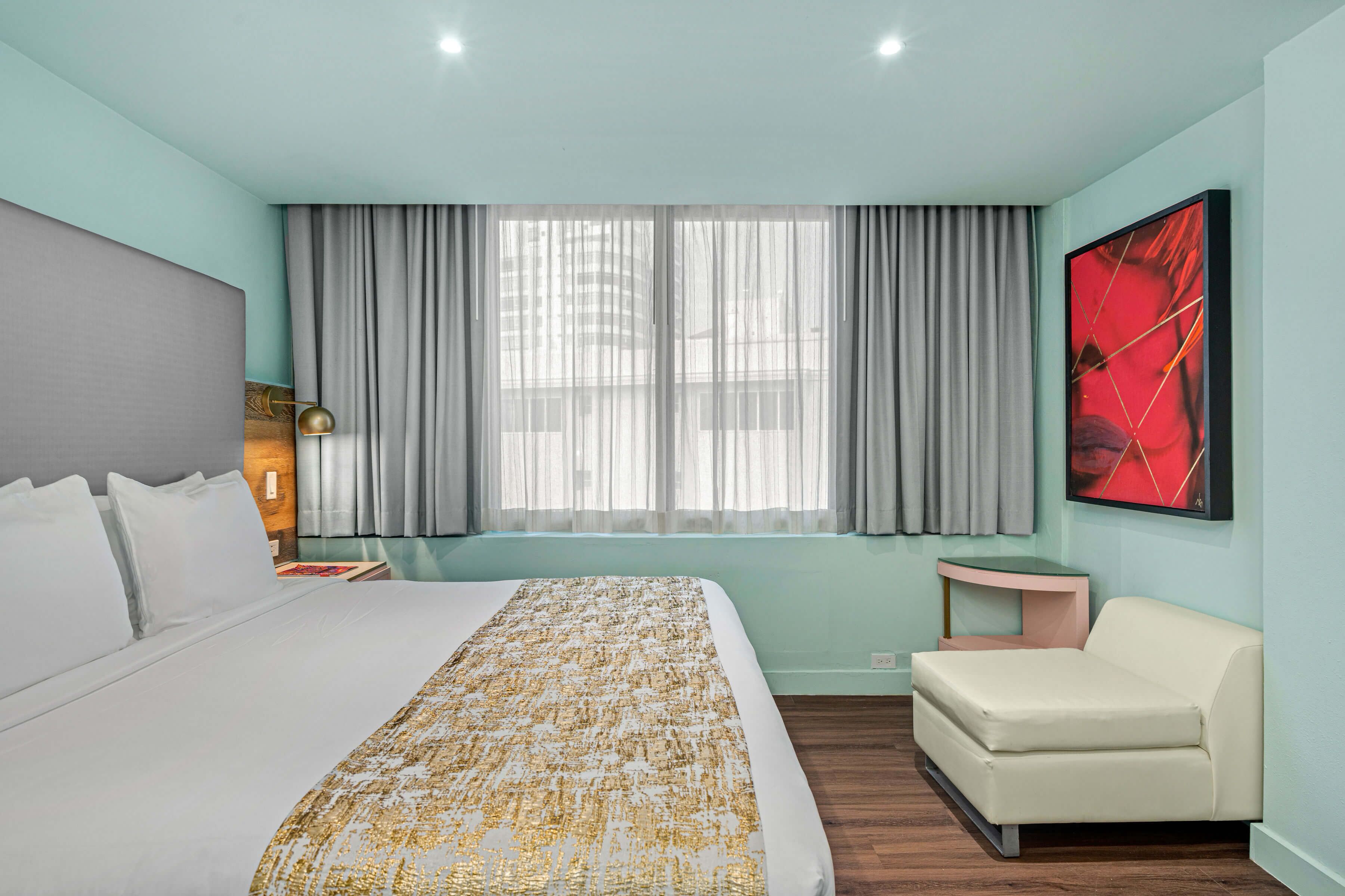 A hotel room with a large bed, a bedside table with a lamp, a small sofa, a window with curtains, and a red abstract painting on the wall.