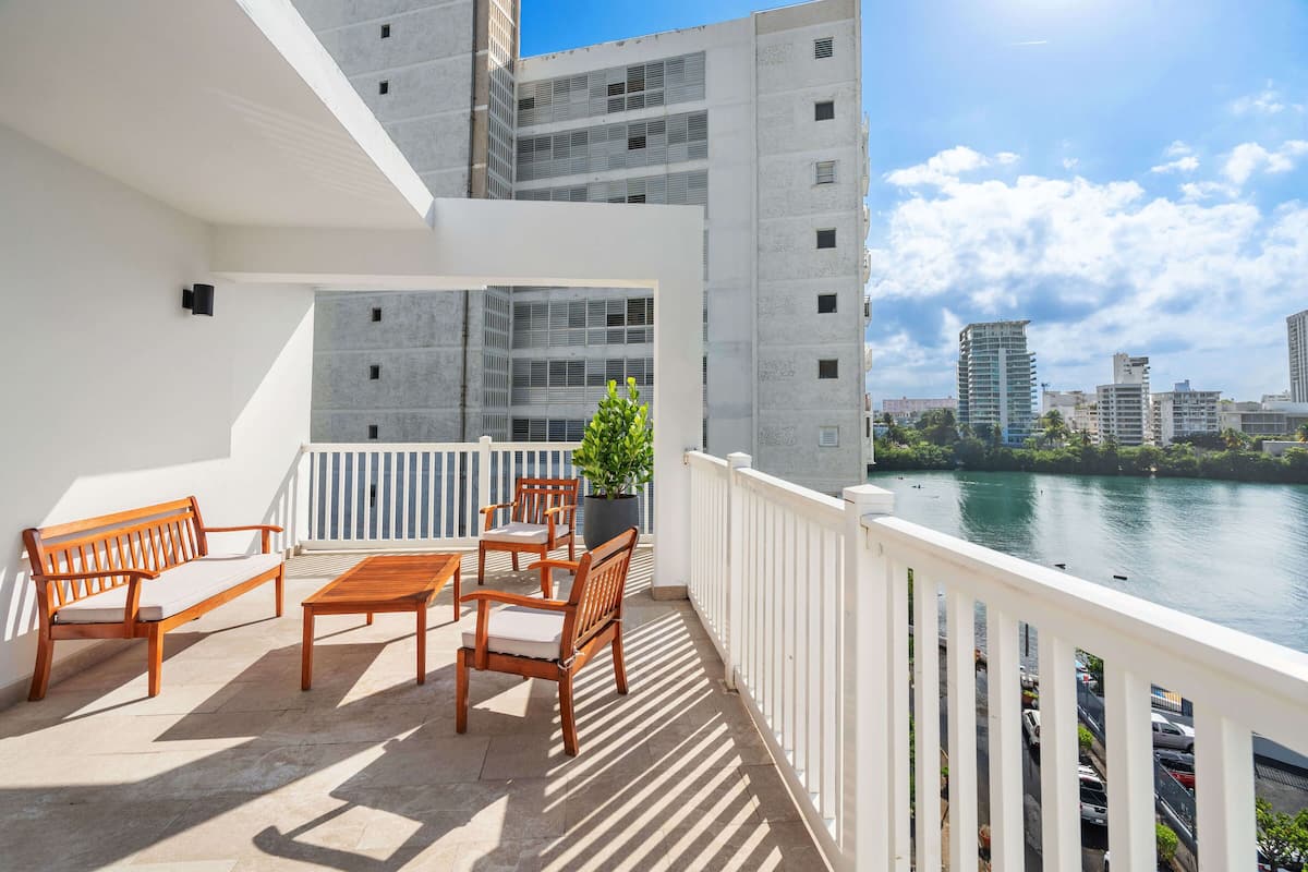 A balcony with a wooden bench, a table, and chairs, overlooking a river with buildings in the background.