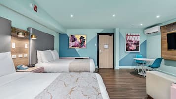 A modern hotel room with a bed, bedside table, lamp, artwork, and a small dining area with a table and chairs.