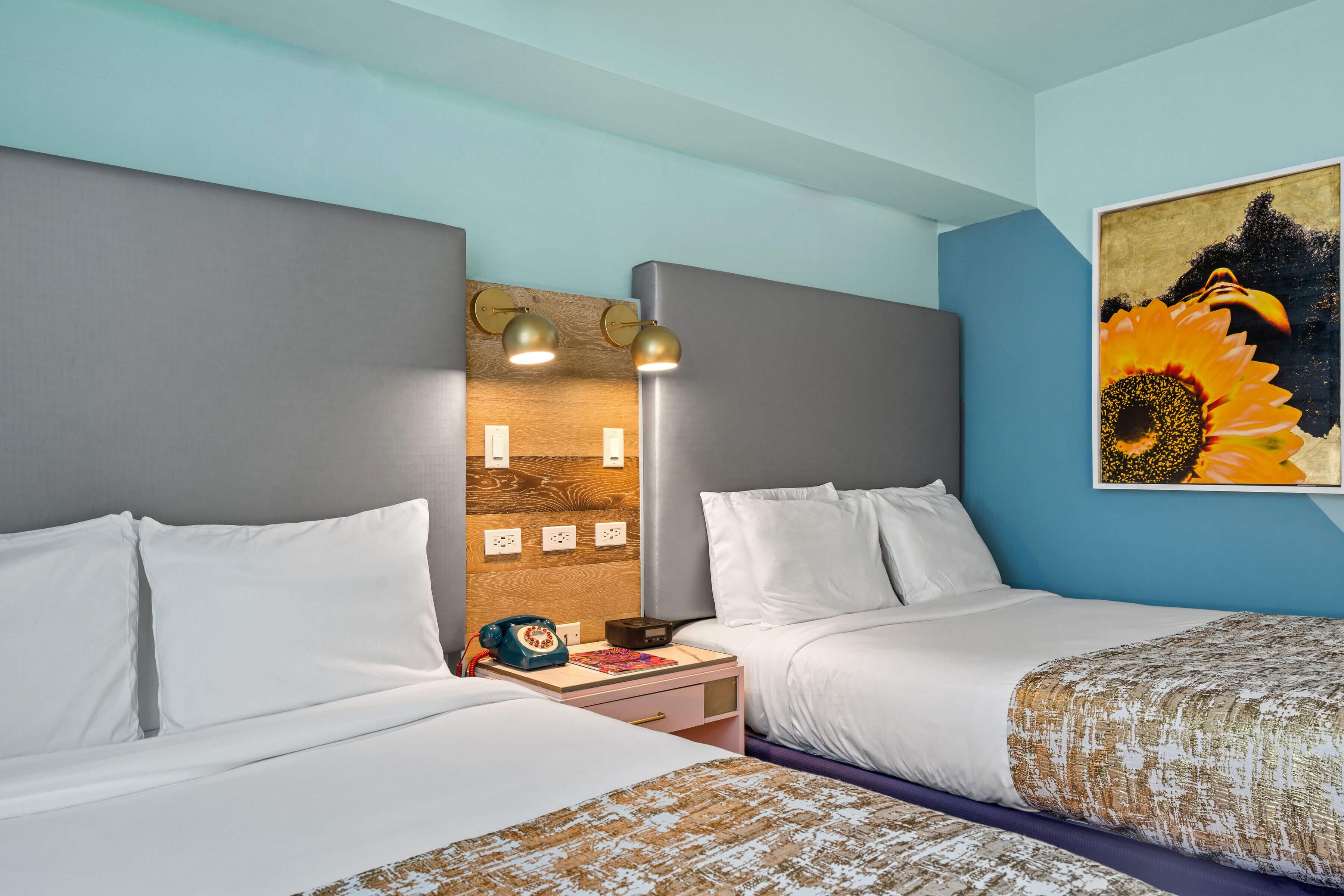 A hotel room with two beds, a nightstand, a lamp, and a painting of a sunflower.