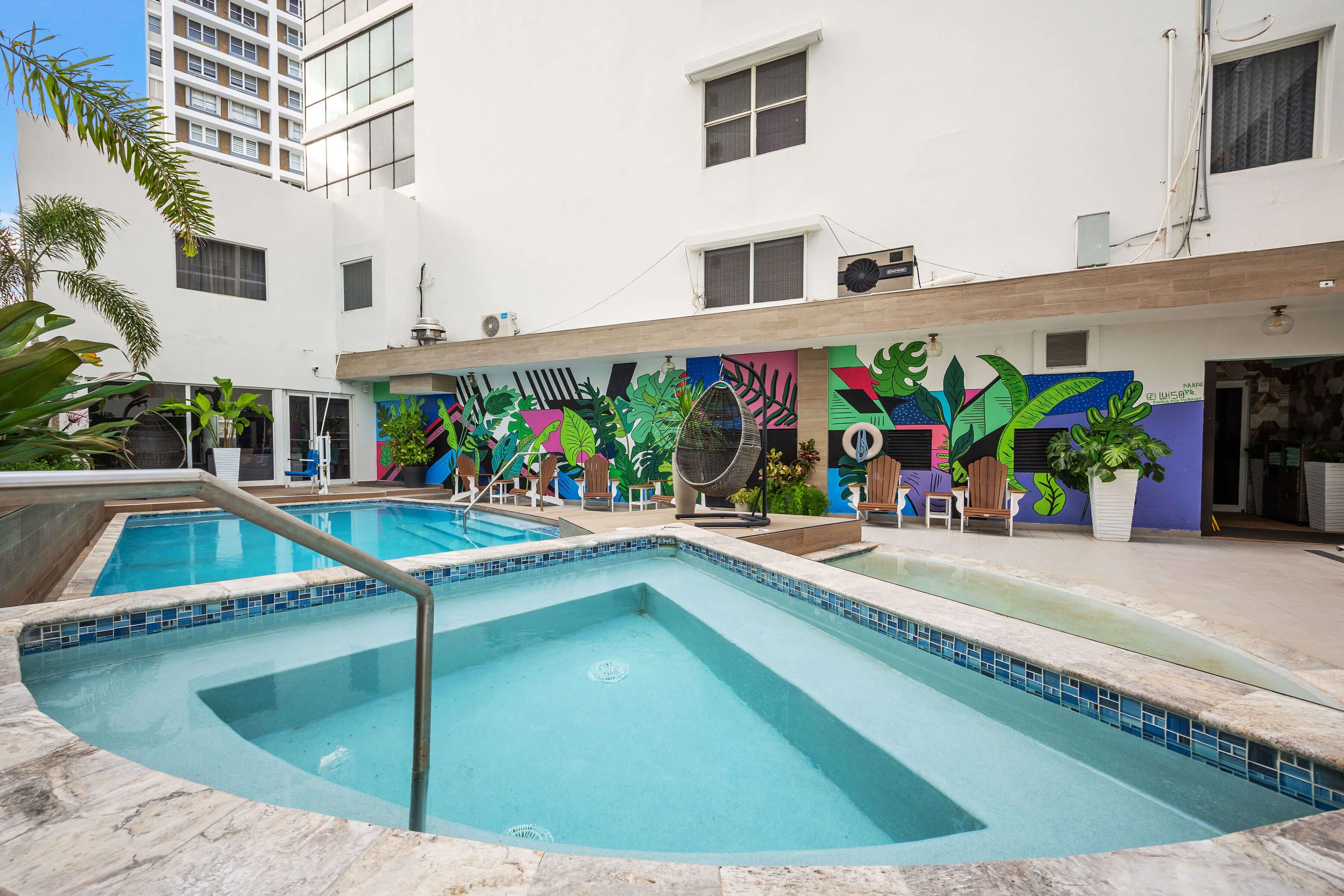 Une piscine dont le bord est recouvert de carreaux, entourée d’un bâtiment blanc orné d’une fresque colorée.
