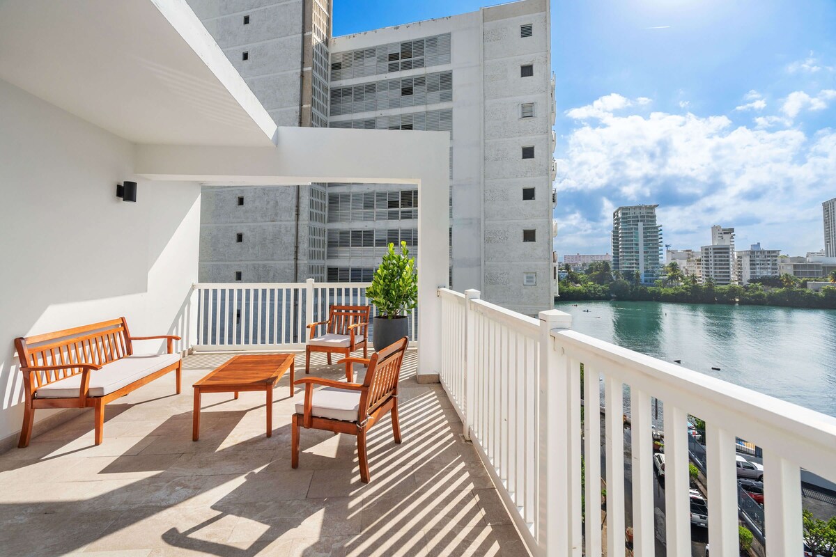 A balcony with a wooden bench, a table, and chairs, overlooking a river with buildings in the background.