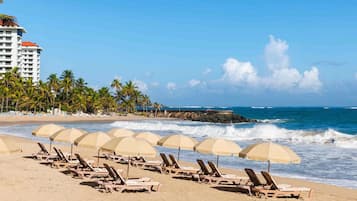 Una playa con filas de tumbonas y sombrillas, un hotel al fondo y un espigón rocoso.
