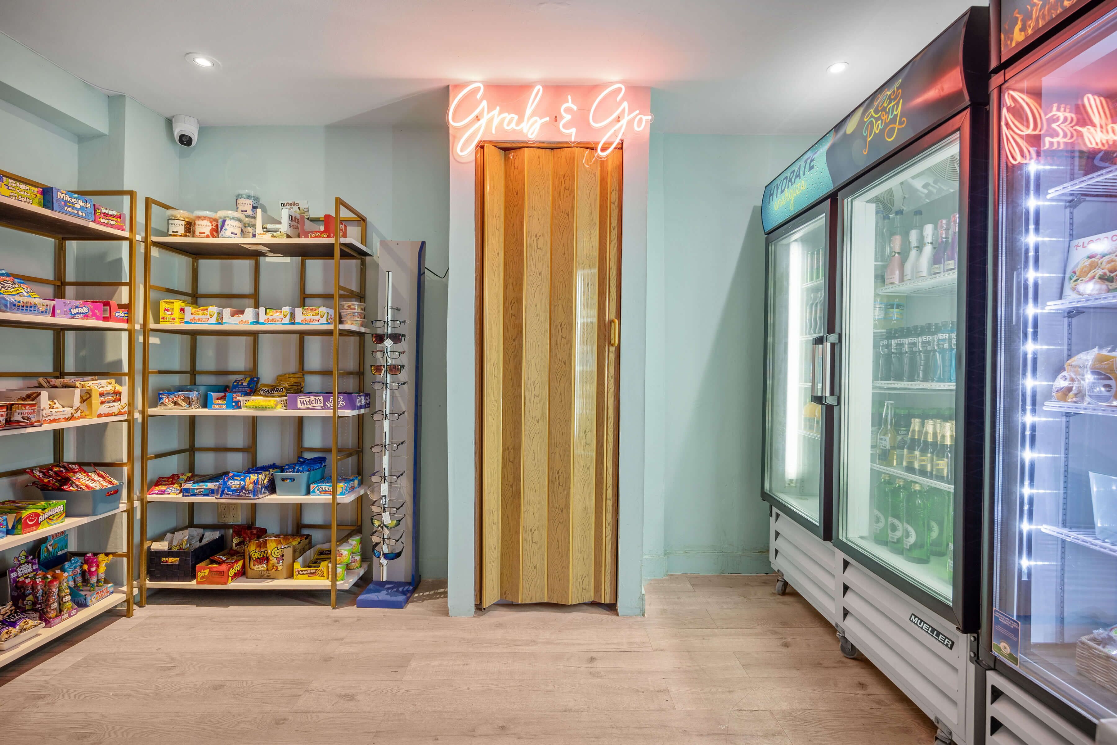 A store interior with shelves stocked with snacks and beverages, a neon sign reading "Grab & Go", and a refrigerated display case.