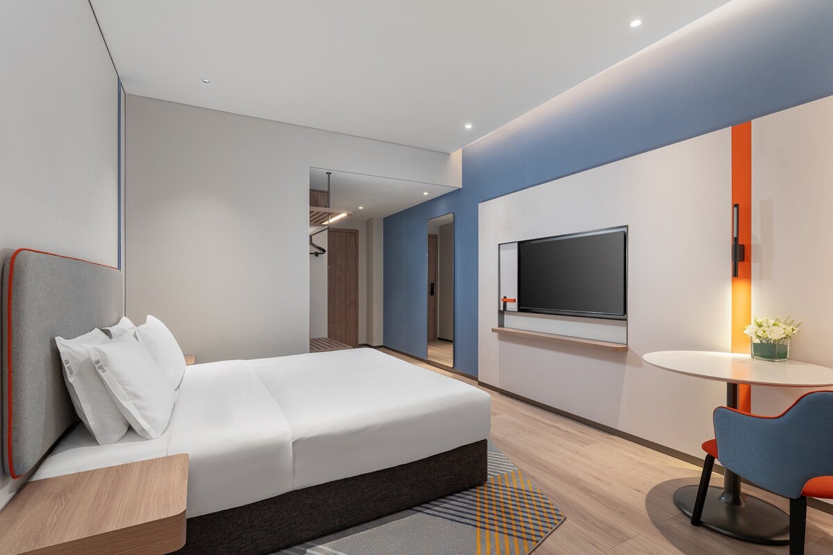 Una habitación de hotel moderna con una cama, un televisor, una pequeña mesa con una silla y un baño a la vista al fondo.
