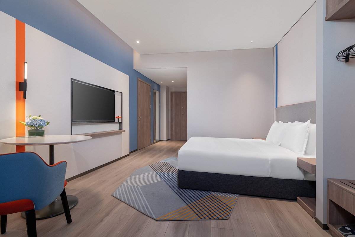 Una habitación de hotel moderna con una cama, un televisor, una mesita con una silla y un jarrón decorativo.