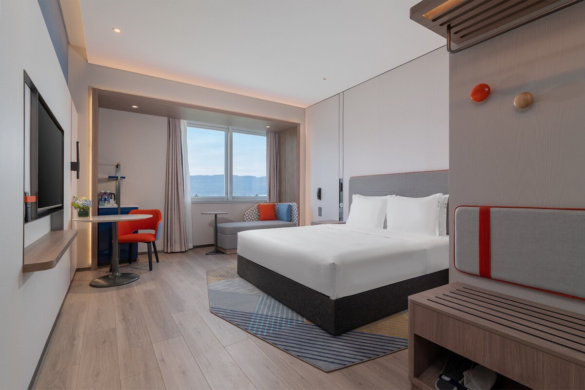 Habitación de hotel moderna con una cama grande, un escritorio con silla, un pequeño rincón de descanso y vistas al exterior.