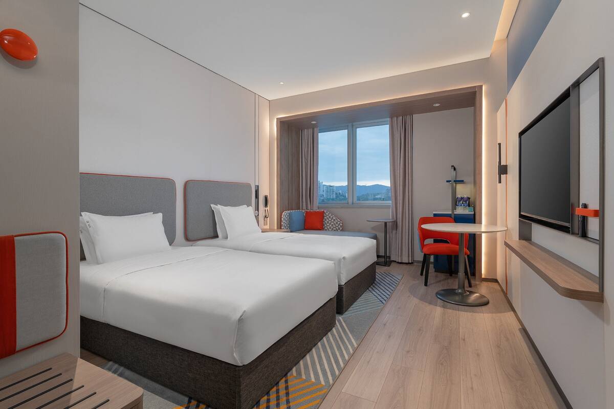 Habitación de hotel con dos camas, un escritorio, una silla, un televisor y una ventana con vistas a la ciudad.