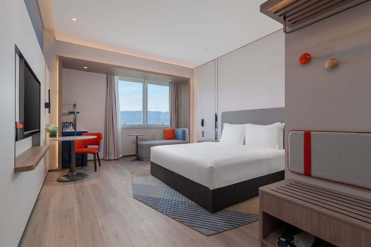 Habitación de hotel moderna con una cama grande, un escritorio con silla, un pequeño rincón de descanso y vistas al exterior.
