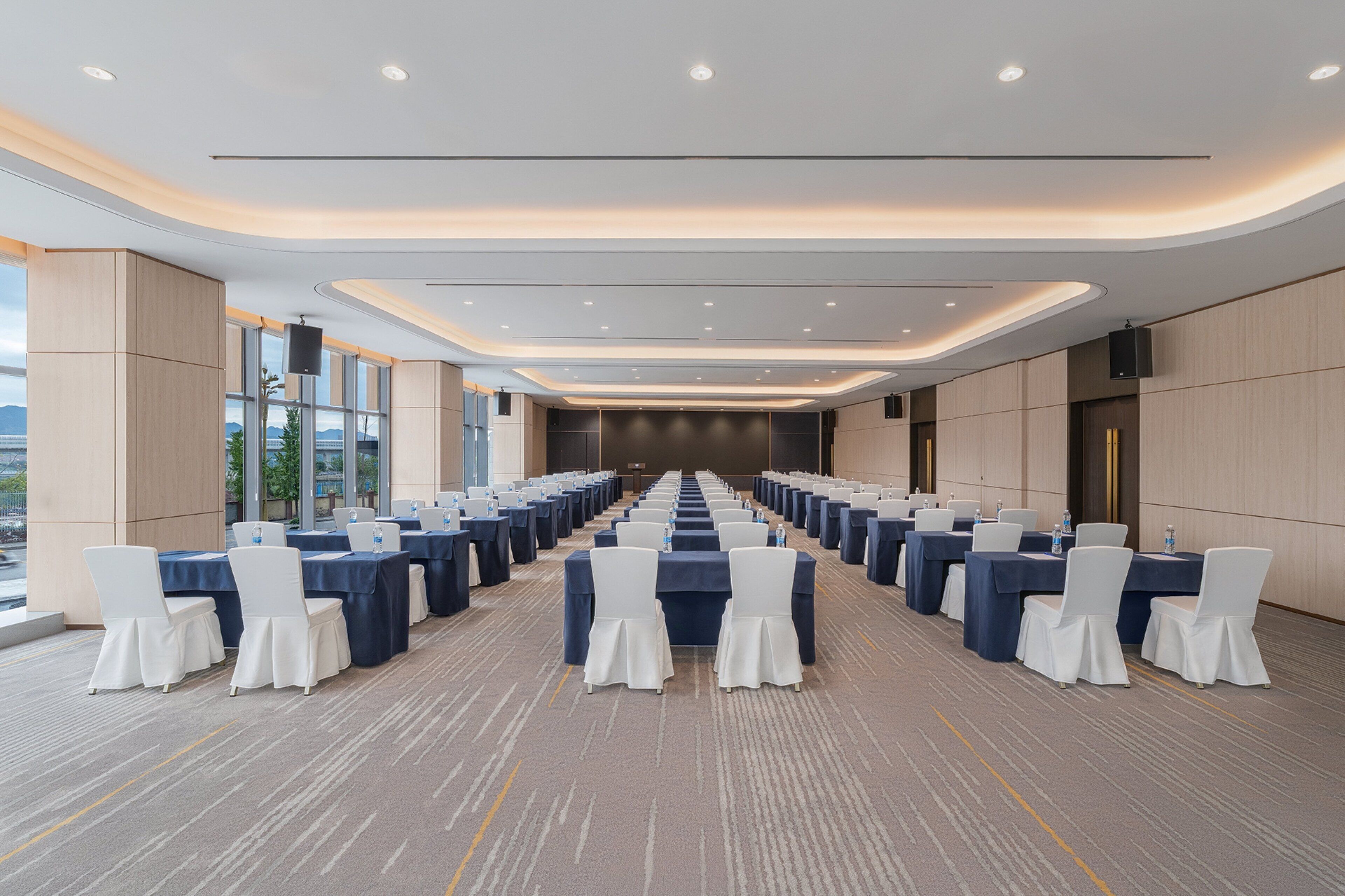 Un salón de conferencias con filas de mesas y sillas, ventanas amplias y un diseño moderno del techo.