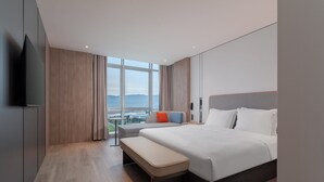 Ett modernt hotellrum med en stor säng, en bänk, en tv och utsikt över staden genom stora fönster.