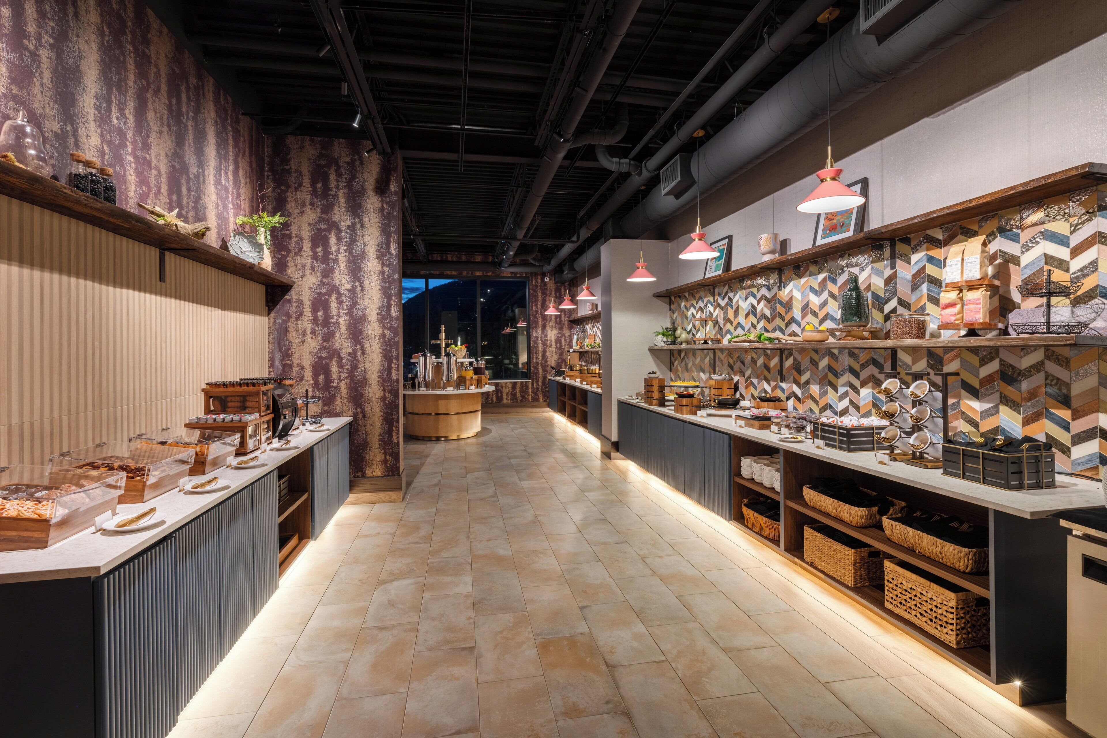 A modern cafe with a long counter displaying various food items, a rustic wall with a herringbone pattern, and pendant lighting.