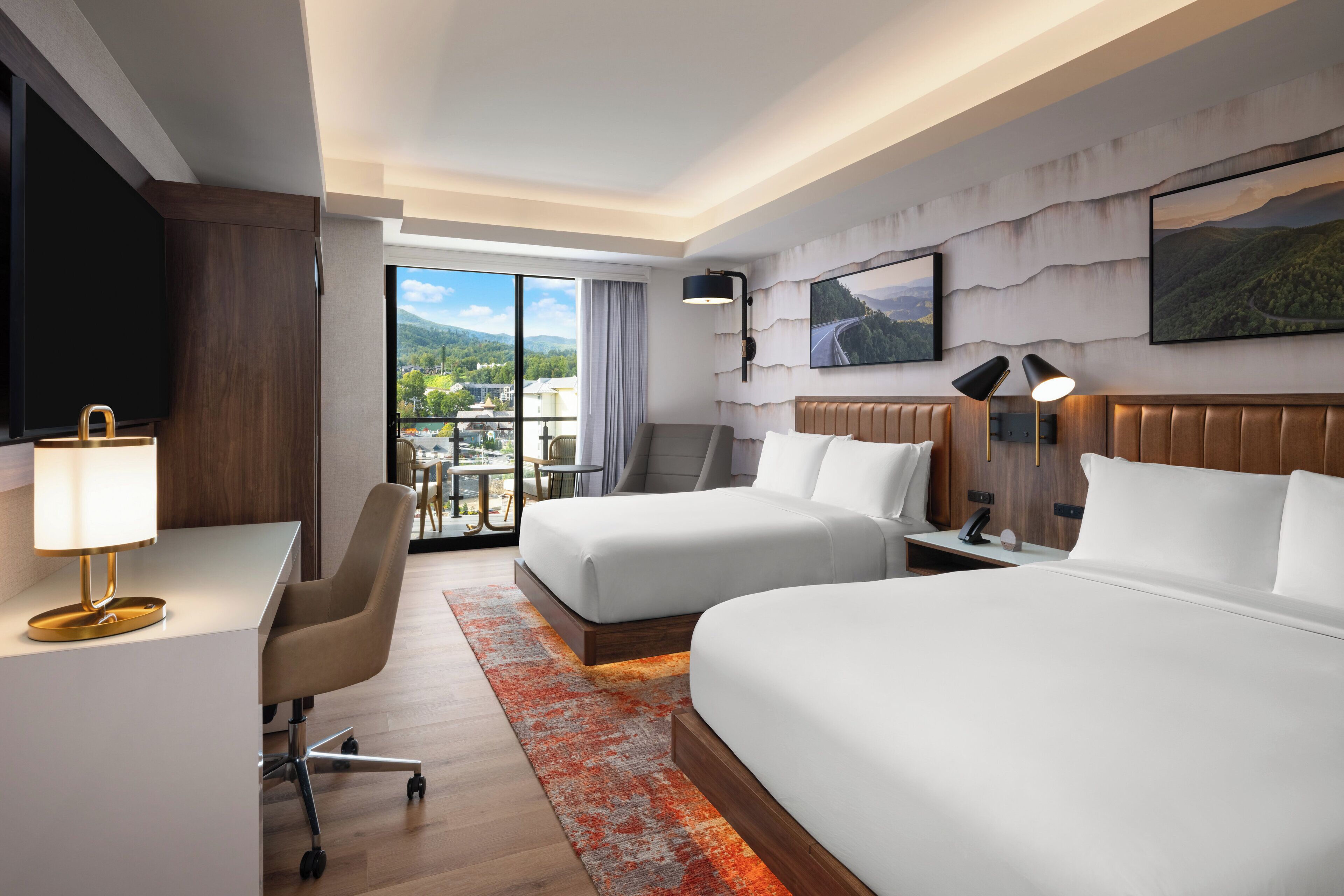 A hotel room with two beds, a desk, a chair, a TV, and a balcony with a view of the mountains.
