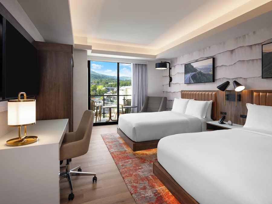 A hotel room with two beds, a desk, a chair, a TV, and a balcony with a view of the mountains.