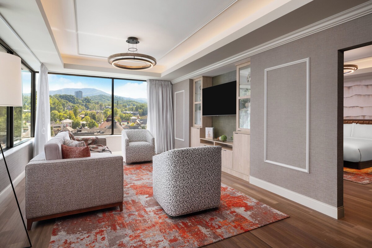 A modern living room with a sofa, ottoman, and TV, large windows with a city view, and a fireplace.