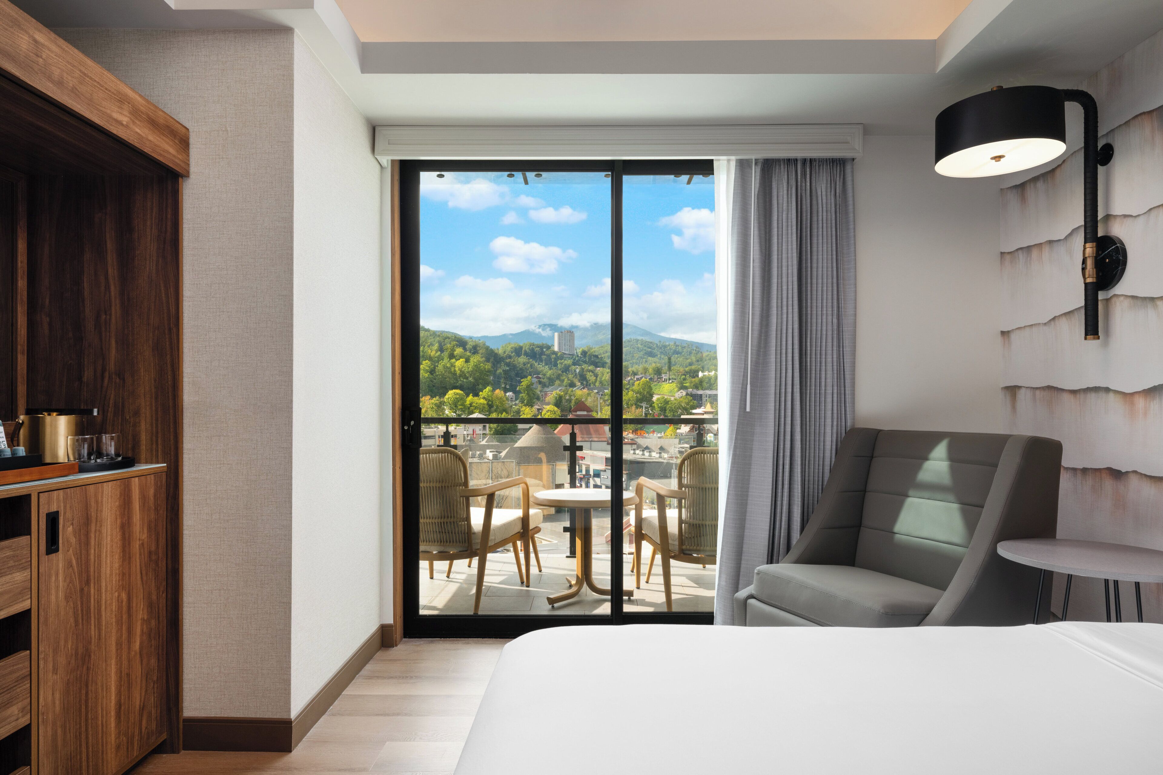 A hotel room with a large bed, a wooden cabinet, a dining area with chairs, a table, and a view of a cityscape through a sliding door.