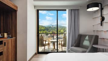 A hotel room with a large bed, a wooden cabinet, a dining area with chairs, a table, and a view of a cityscape through a sliding door.