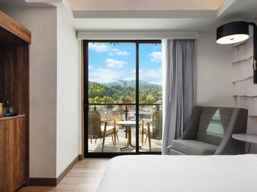 A hotel room with a large bed, a wooden cabinet, a dining area with chairs, a table, and a view of a cityscape through a sliding door.