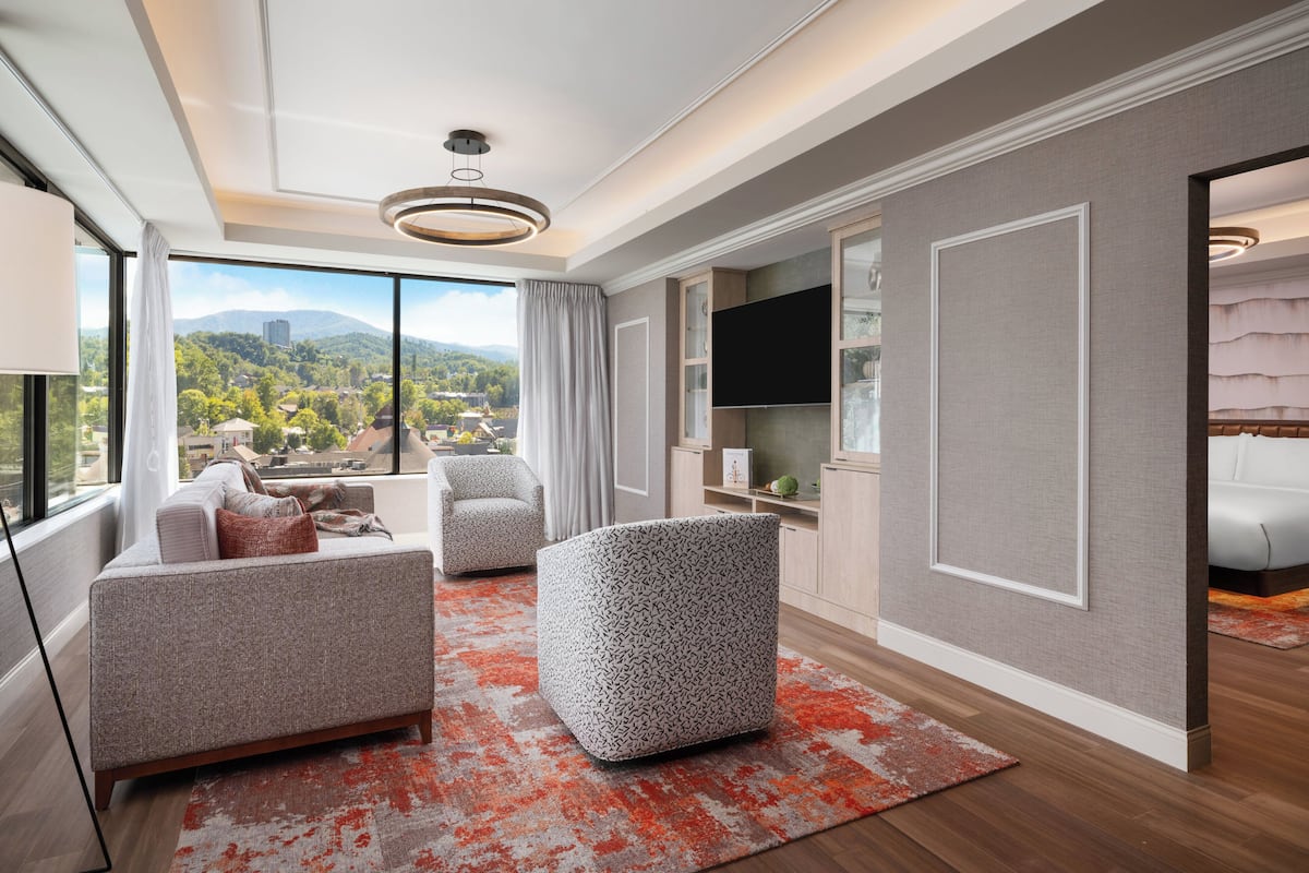 A modern living room with a sofa, ottoman, and TV, large windows with a city view, and a fireplace.