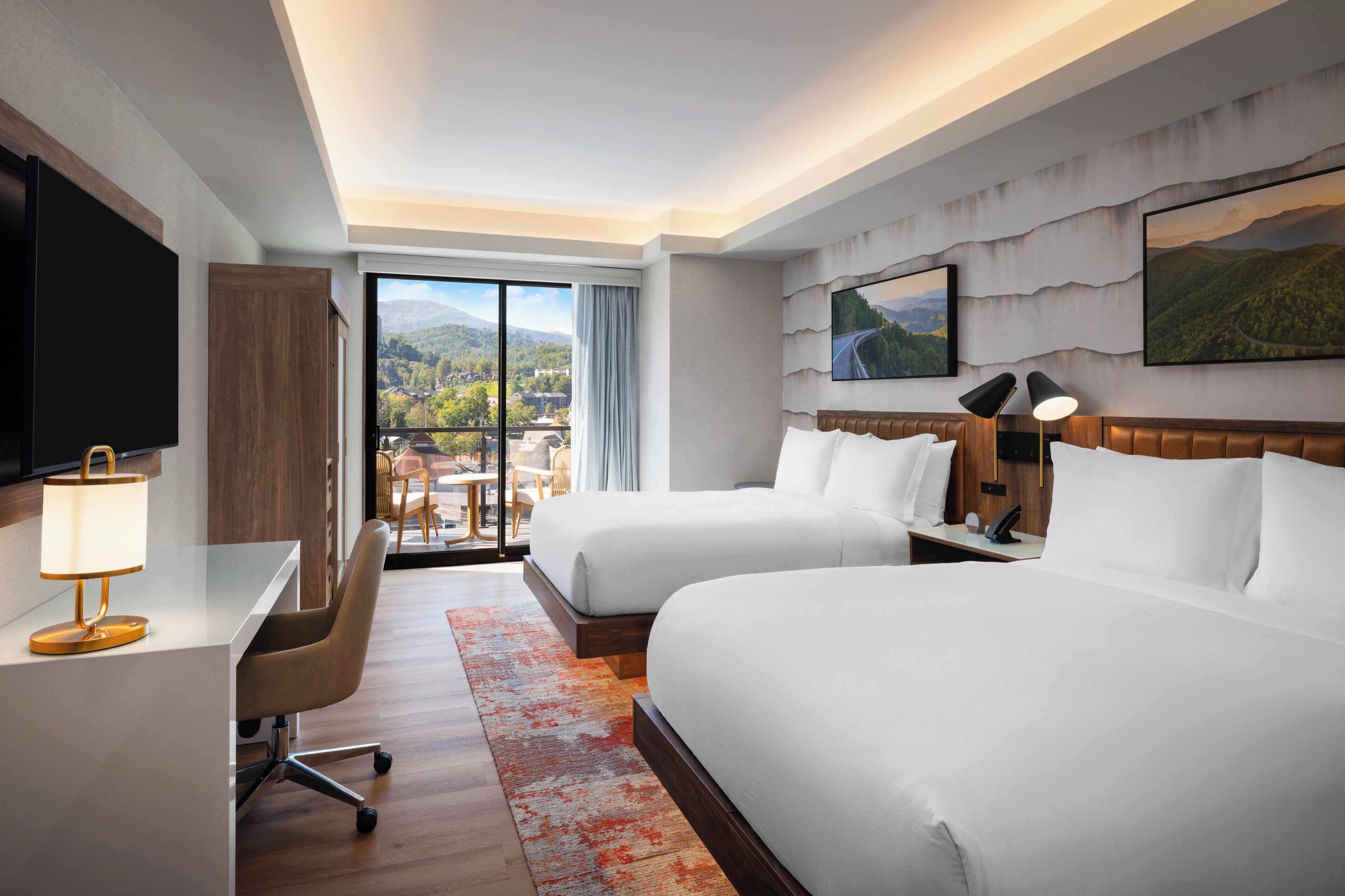 A hotel room with two beds, a desk, a chair, a TV, and a large window with a mountain view.