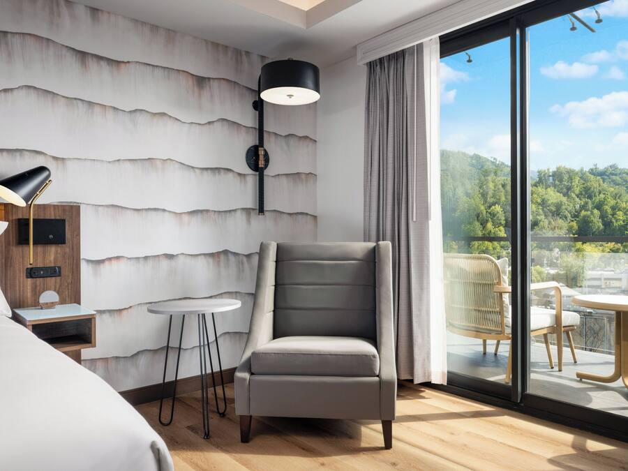 Quarto de hotel moderno com uma cama grande, uma poltrona cinza, uma mesa redonda pequena, uma luminária e vista para uma varanda com mobiliário.
