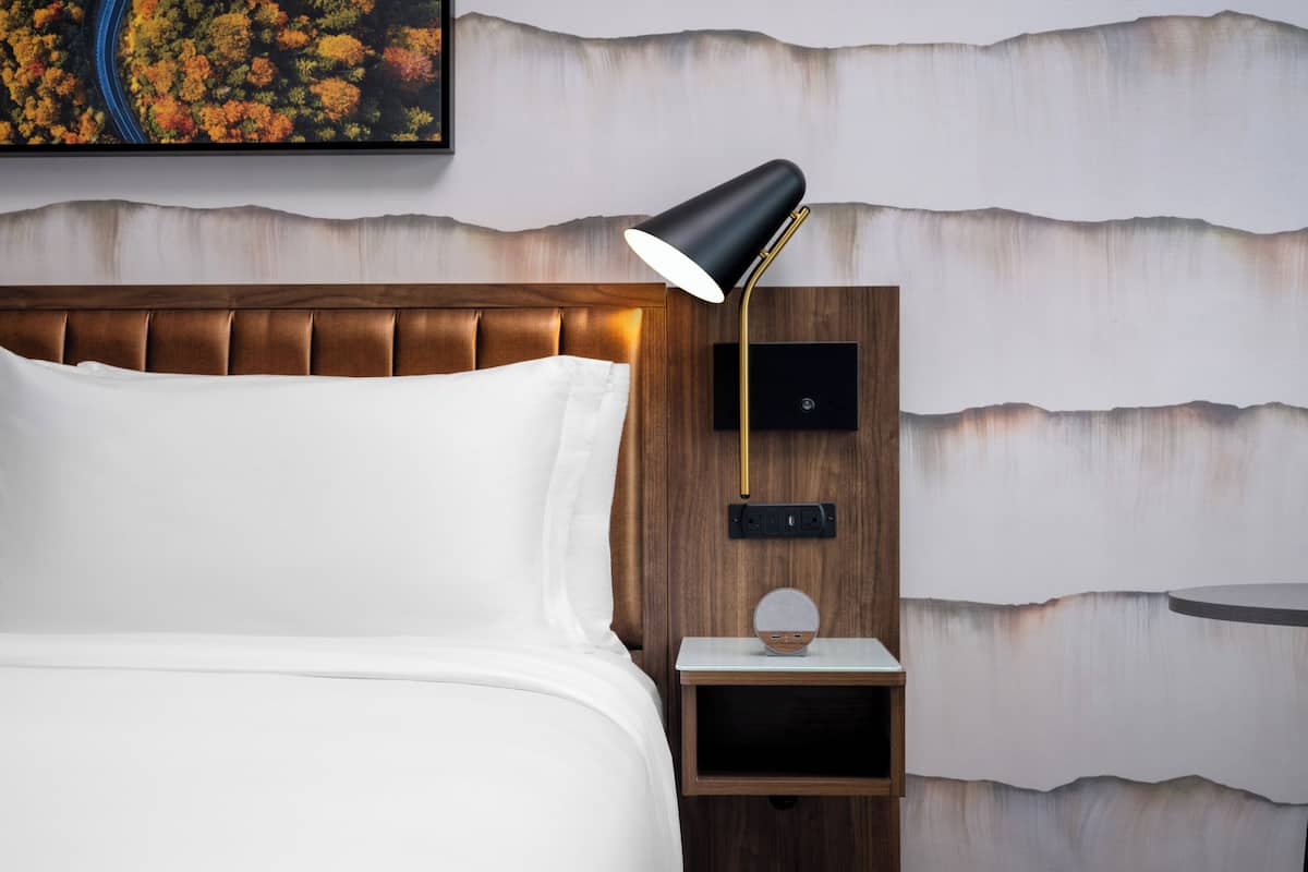 A neatly made bed with white linens, a wooden headboard, a bedside table with a lamp, and a wall-mounted power outlet.