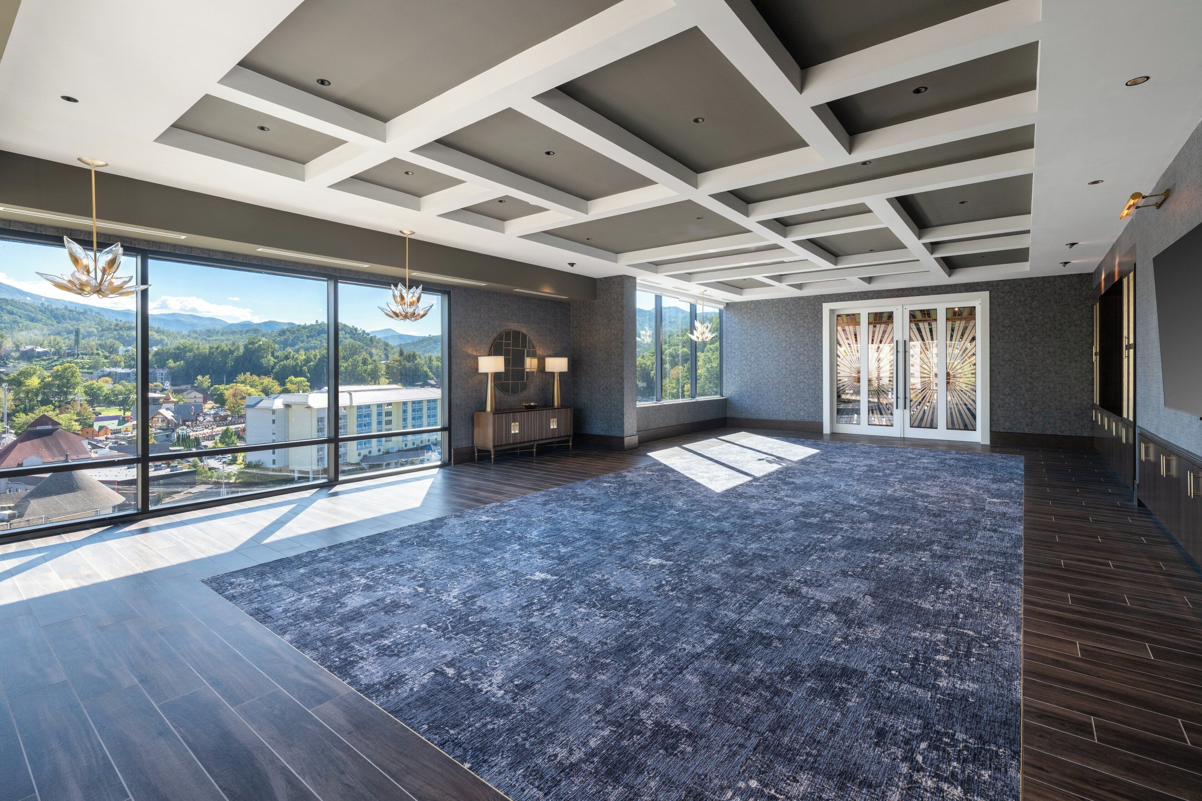 A spacious room with large windows offering a view of a cityscape and mountains. The room features a modern ceiling design with recessed lighting and a dark wood floor.