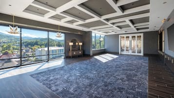 A spacious room with large windows offering a view of a cityscape and mountains. The room features a modern ceiling design with recessed lighting and a dark wood floor.