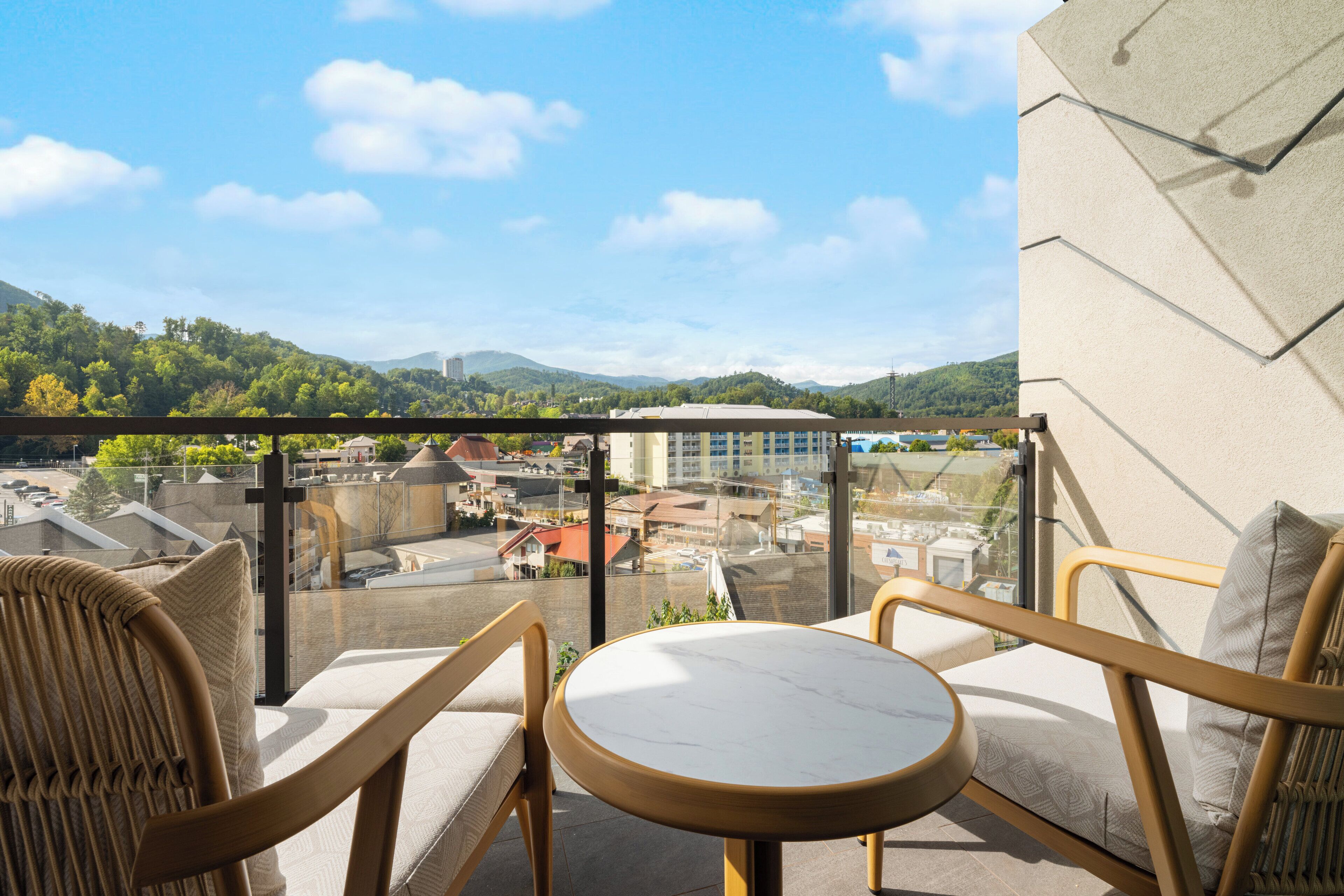 A balcony with wicker chairs and a small round table, overlooking a cityscape with buildings and greenery.