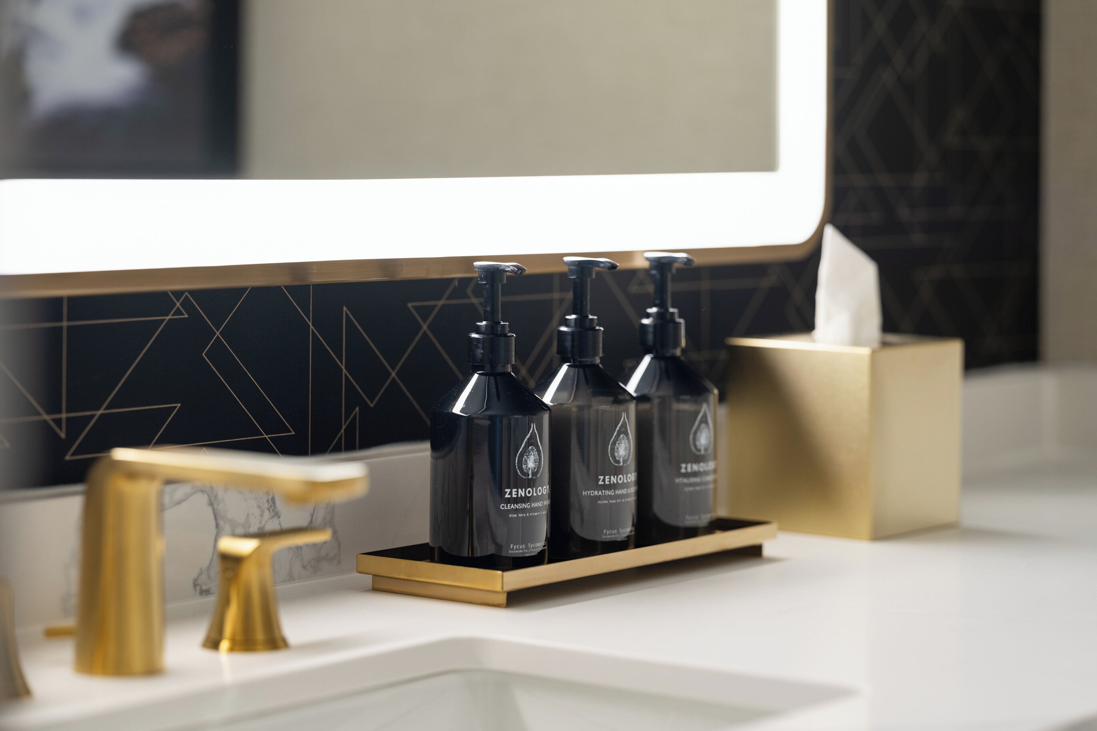 A bathroom vanity with a gold faucet, a white sink, and a black soap dispenser holder with three bottles.