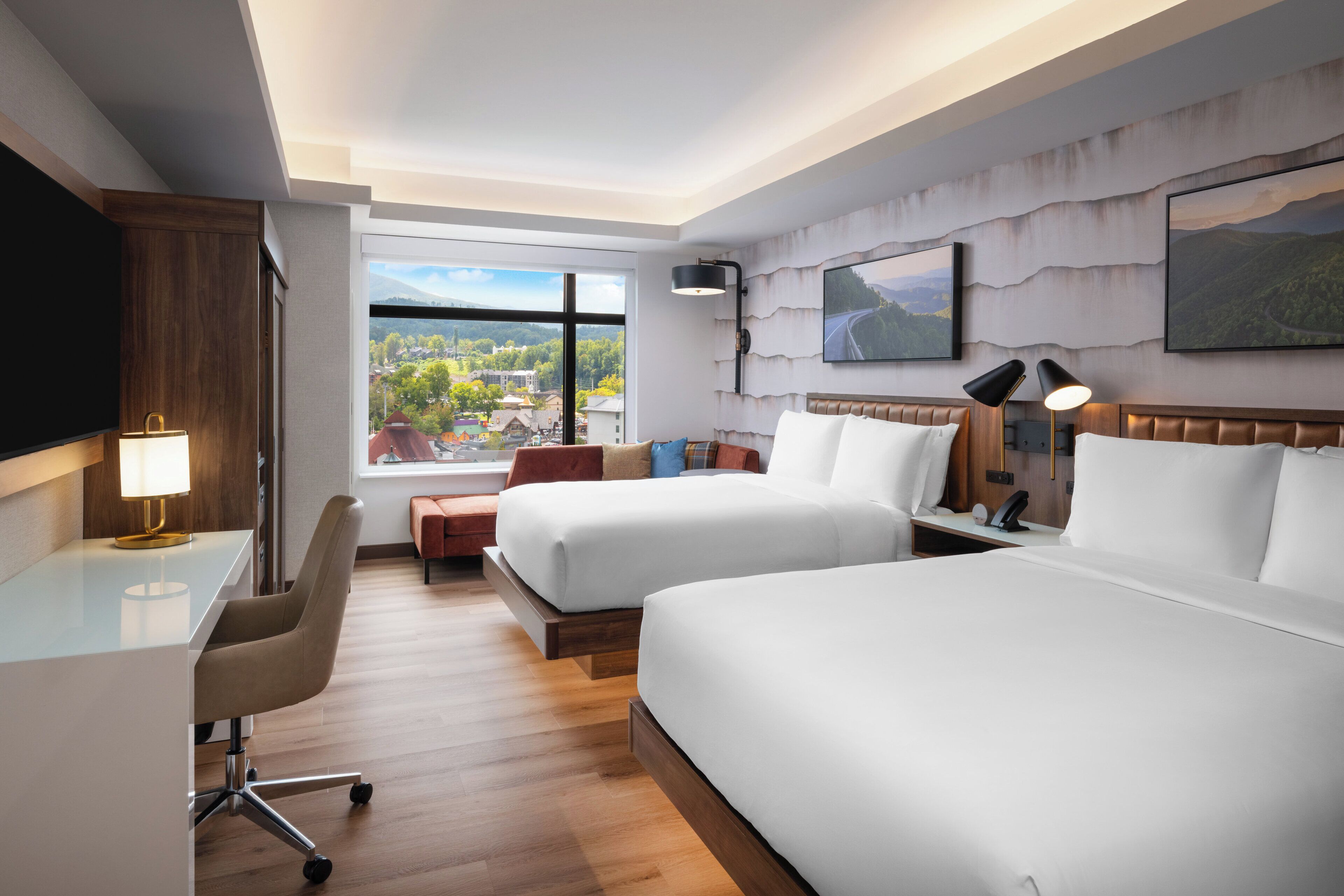 A hotel room with two beds, a desk, a chair, and a large window offering a scenic view.