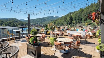 Una terraza en la azotea con varias zonas de estar, incluyendo mesas y sillas redondas, rodeada de macetas con plantas y flores.