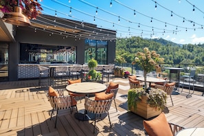 A rooftop bar with outdoor seating, a bar area, and a view of a forested hill.