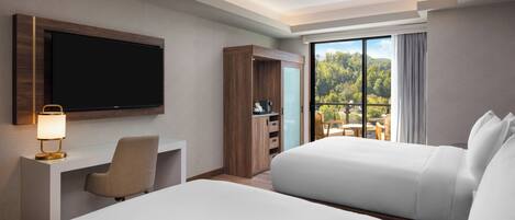 Habitación de hotel con una cama grande, un escritorio con lámpara, televisión y puerta corrediza de vidrio que da a un balcón con vistas a árboles.