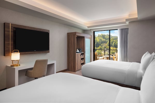 A hotel room with a large bed, a desk with a lamp, a TV, and a sliding glass door leading to a balcony with a view of trees.