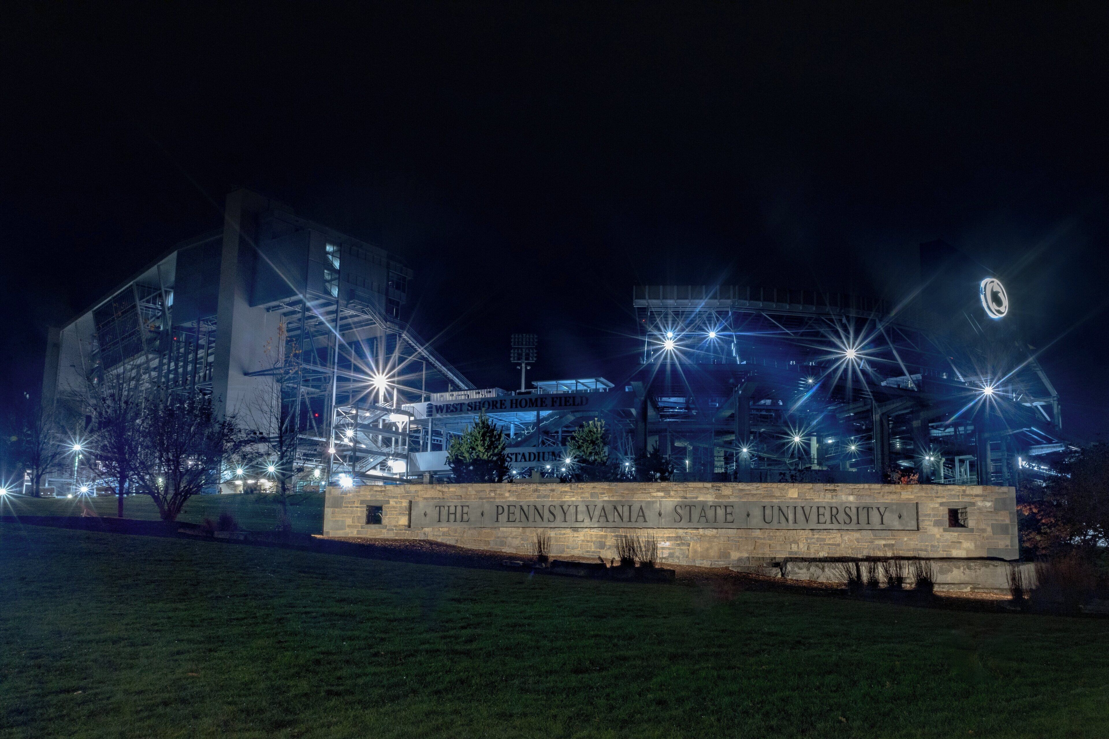 Una vista nocturna del campus de la Universidad Estatal de Pensilvania, con edificios iluminados y un cartel destacando.