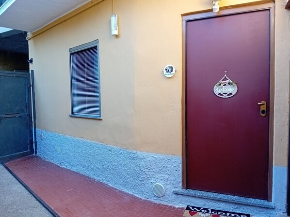 Une porte rouge avec une pancarte « Bienvenue », une petite fenêtre avec des stores et un appareil de climatisation blanc fixé sur un mur beige.