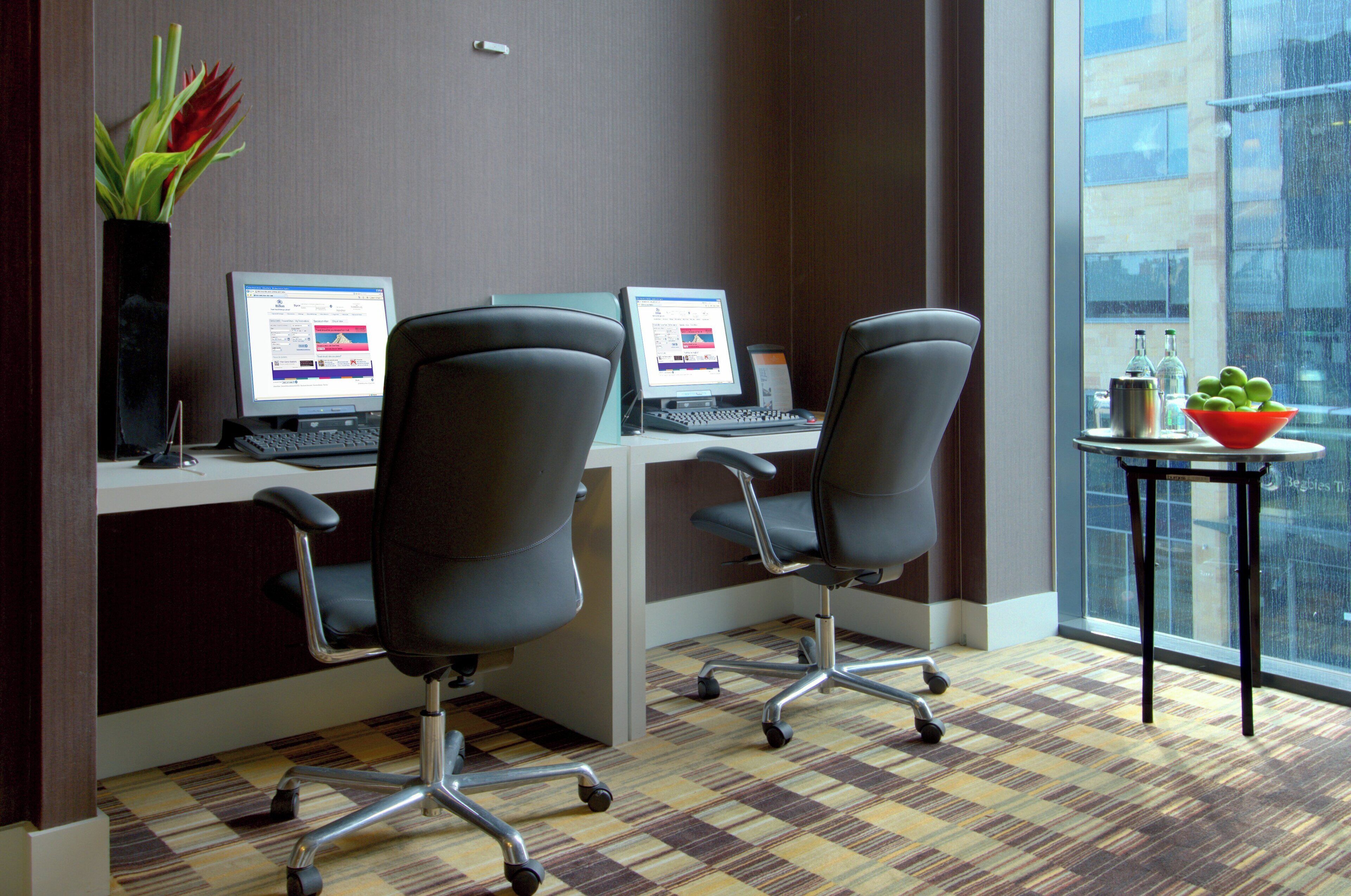 A modern office space with two desks, each with a laptop and an office chair. There is a vase with flowers on the left and a bowl of fruit on the right. The room has a checkered floor and large windows.