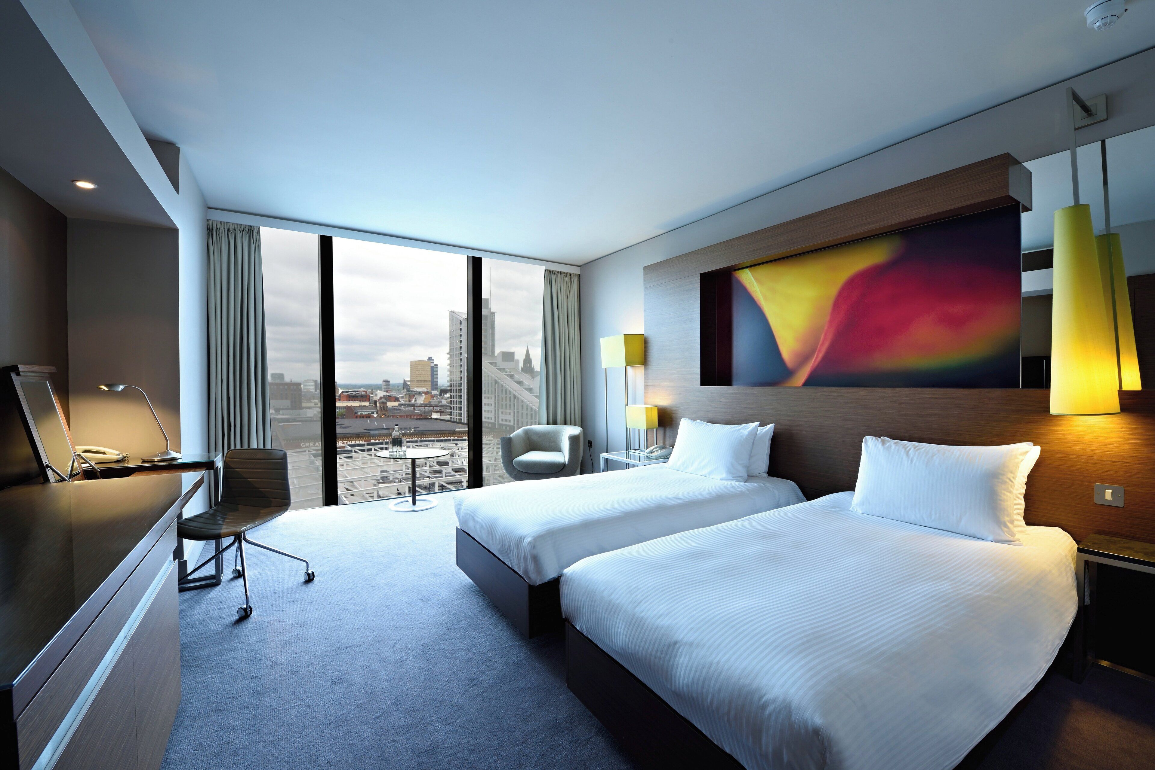 Une chambre d’hôtel avec deux lits, un bureau, une chaise, une grande fenêtre donnant sur la ville et une peinture abstraite aux couleurs vives accrochée au mur.