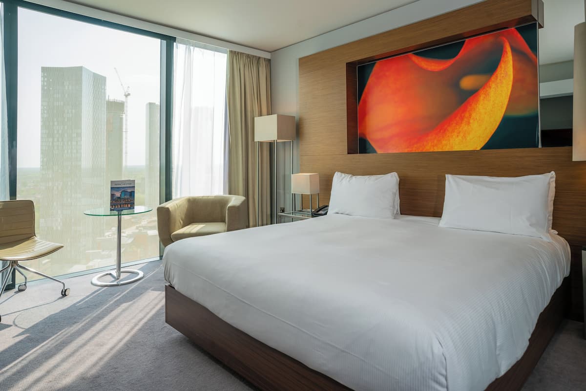 Une chambre d’hôtel avec un grand lit, un bureau, une chaise, une lampe et une vue sur le paysage urbain.