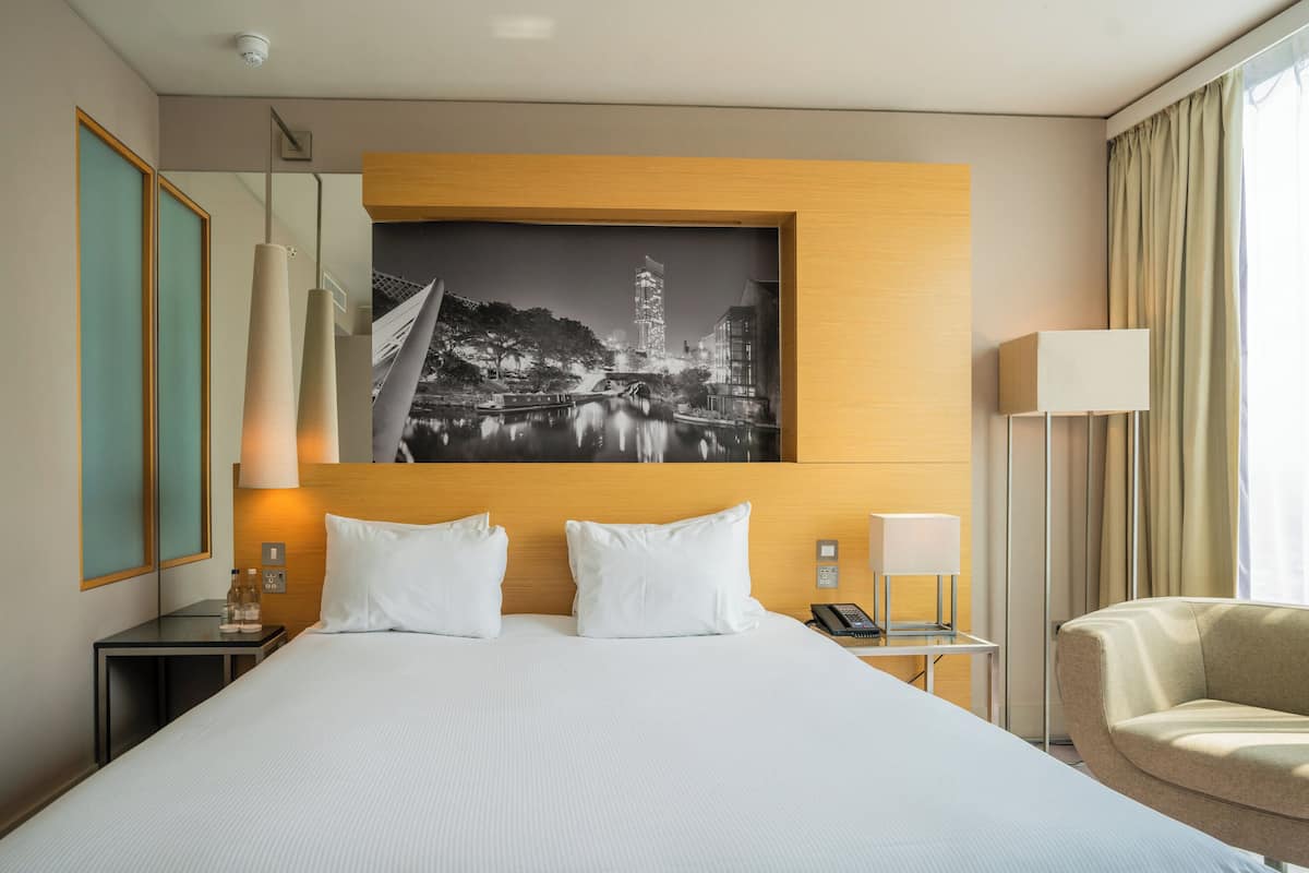 Een hotelkamer met een groot bed, twee nachtkastjes, een stoel, een wandlamp en een lijst met een stadsbeeld.