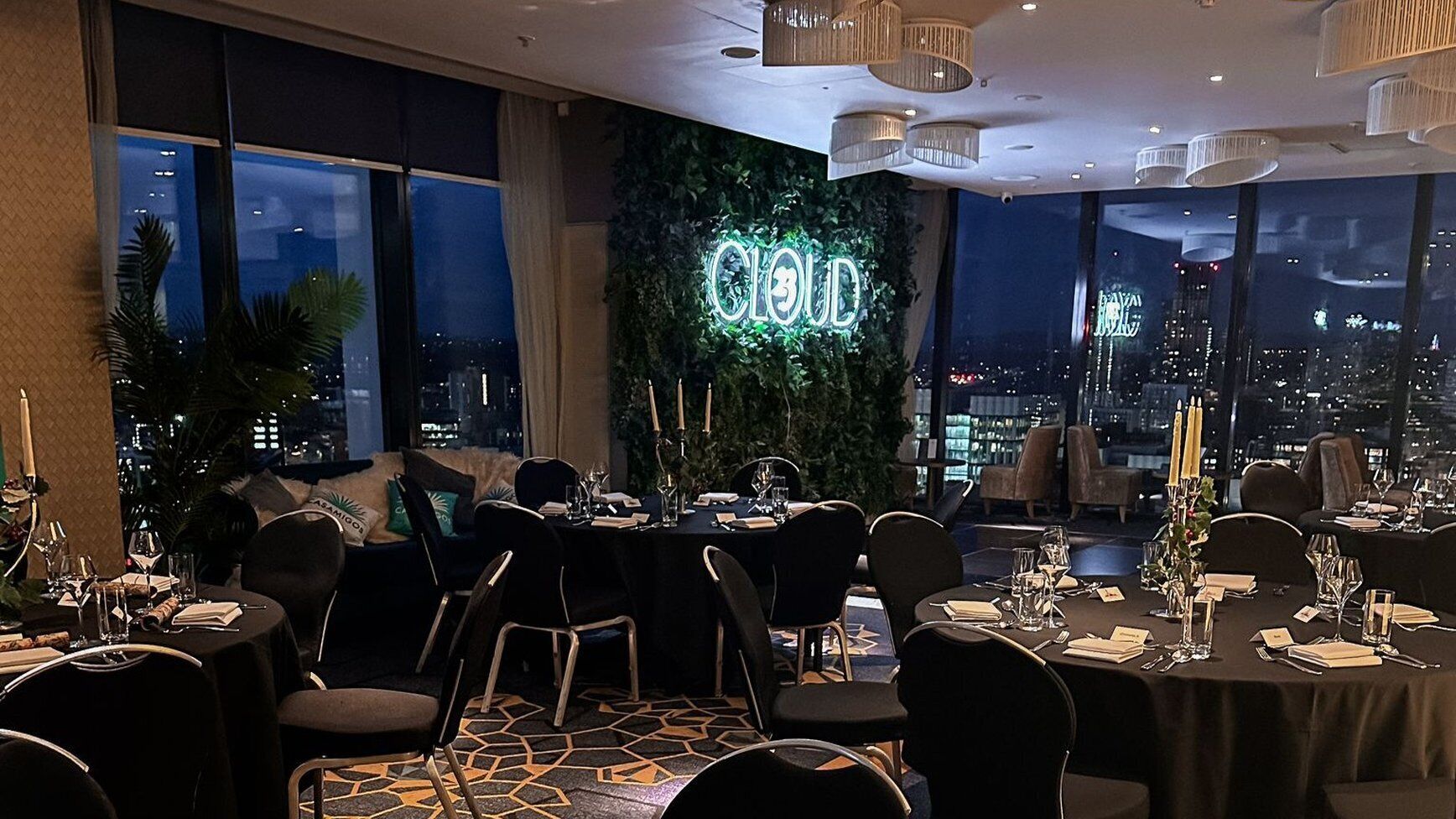 Een restaurant met gedekte tafels, een neonbord waarop 'CLOUD' staat en een stadsgezicht door grote ramen.