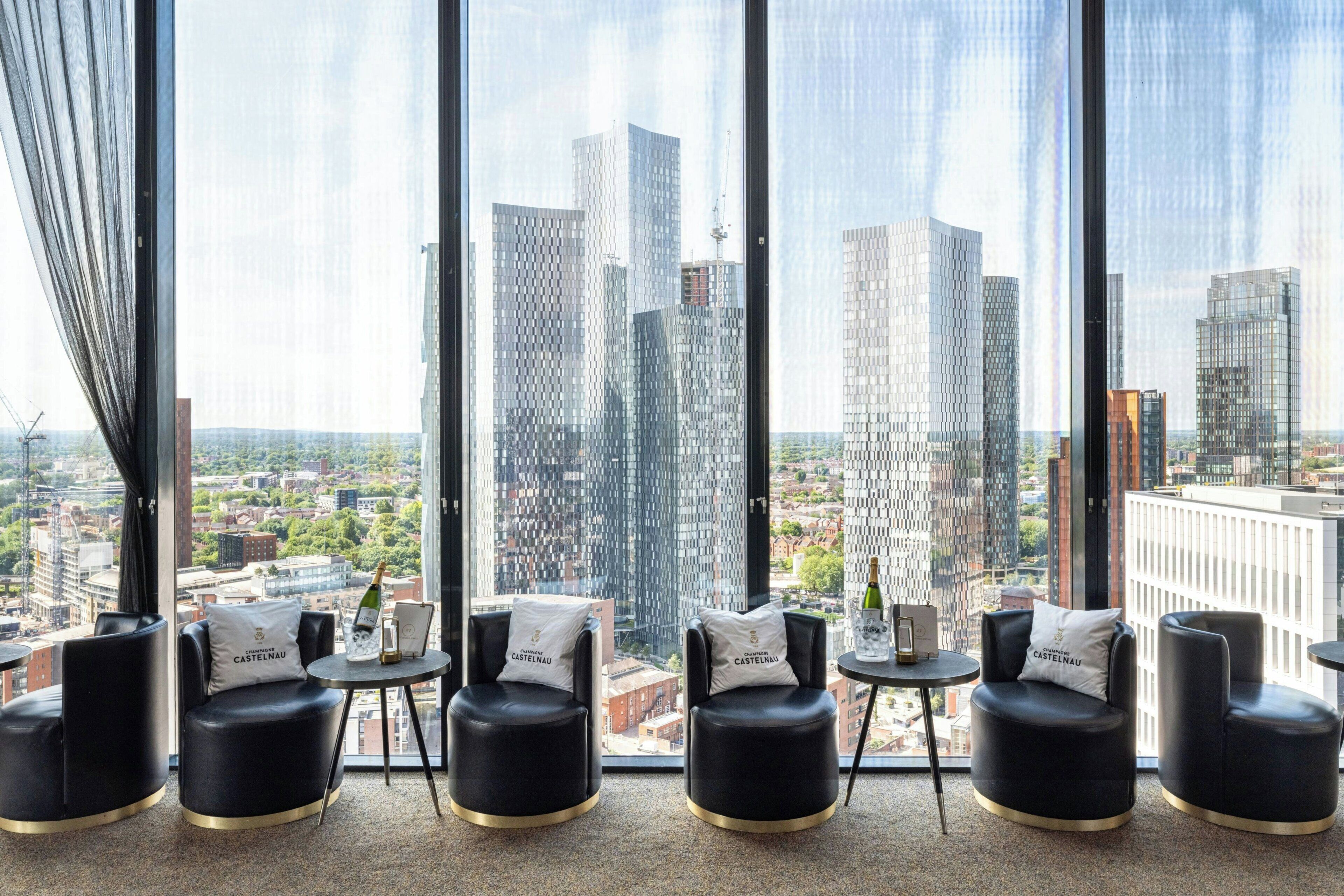 A modern hotel room with large windows, black leather chairs, and a view of the cityscape.